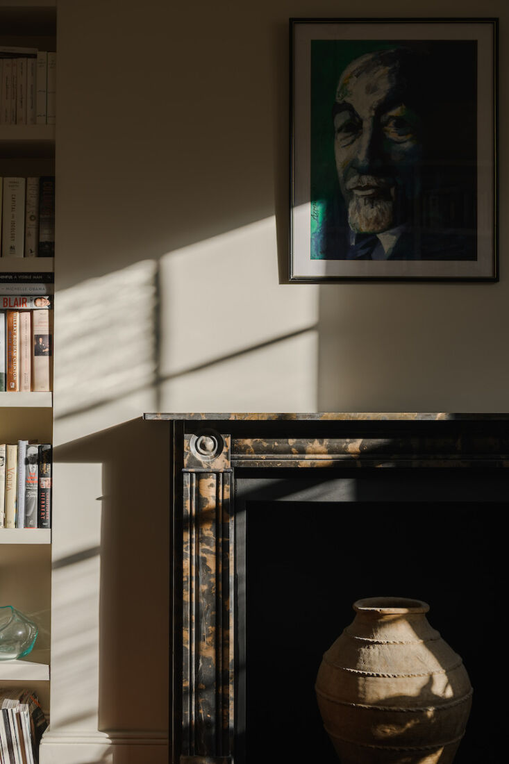 A sunlit study with a traditional marble fireplace, bookshelves, and a portrait.