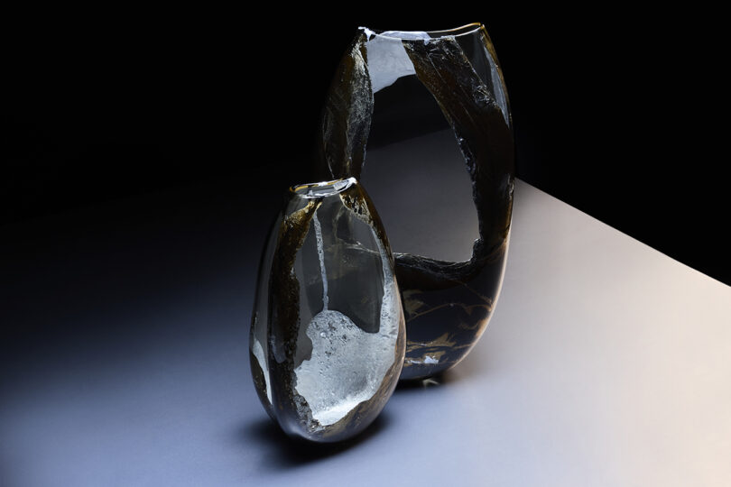 Two sculptural glass objects with metallic inclusions and cracked textures under dramatic lighting.