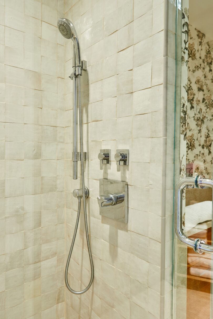 A modern shower with square beige tiles and chrome fixtures, showing a glimpse of floral wallpaper outside.