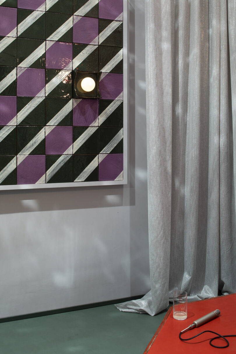 A hospitality setting with a green and purple geometric tile wall, a mounted light, and a red table with a glass.