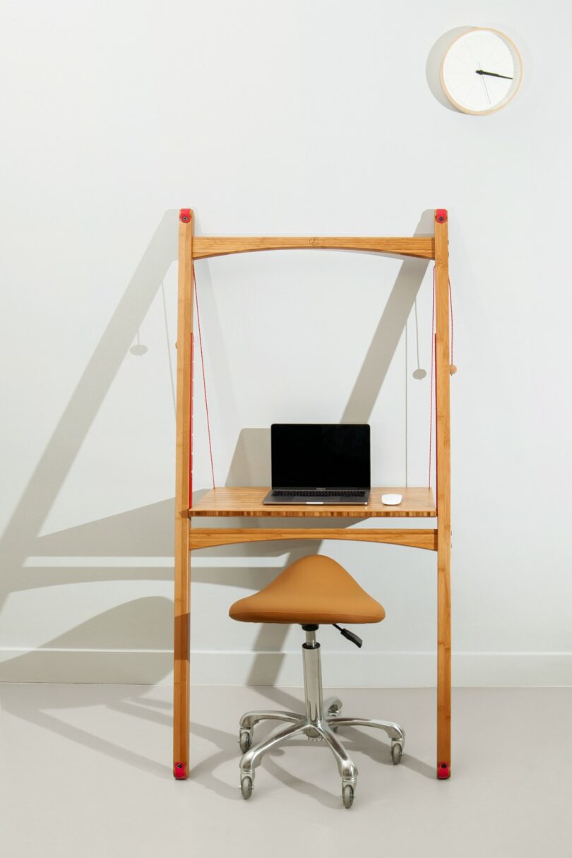 Minimalist wooden standing desk with rope suspension system and a laptop against a white wall.