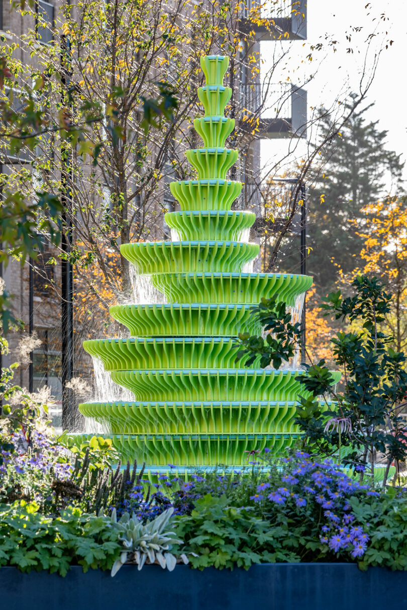 A modern green tiered fountain surrounded by lush garden plants and residential structures.