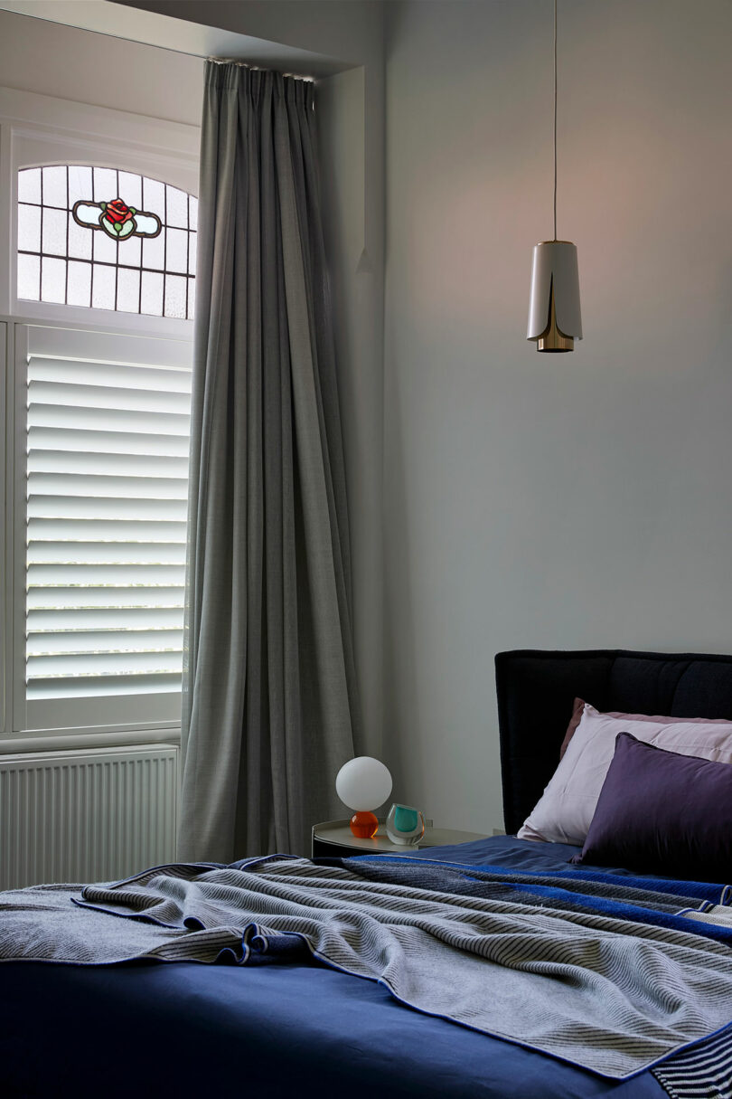 Modern bedroom with blue bedding, white shutters, and a stained glass window above the bed.