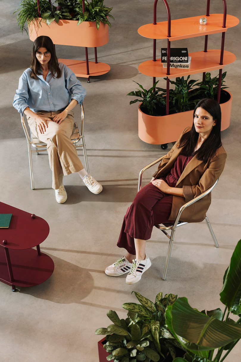 Two women sitting in a modern, plant-filled office lounge area.