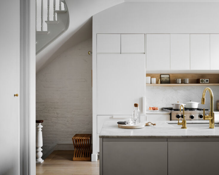 Modern kitchen with white cabinetry, marble countertops, and exposed brick under a staircase.