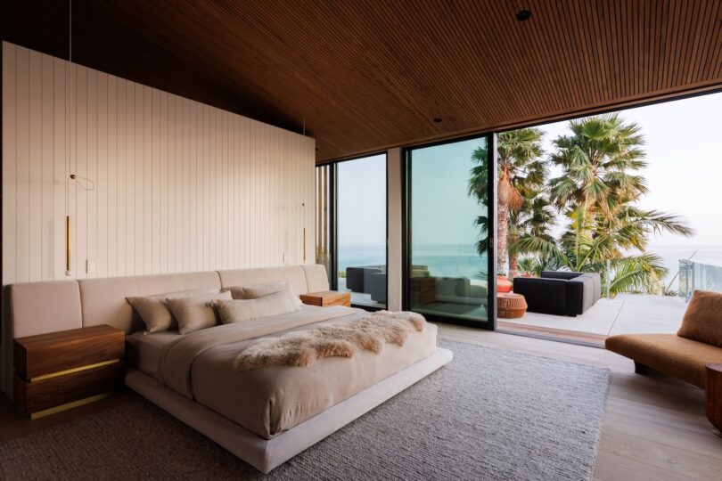 Modern bedroom with wood-paneled ceiling and floor-to-ceiling windows opening to a patio.