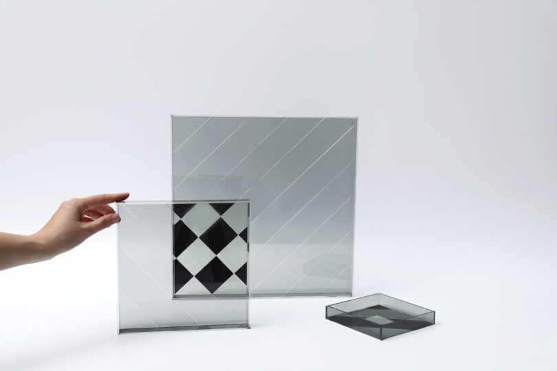A hand holding a square glass pane with a diamond pattern over other glass sheets to show overlapping effects.
