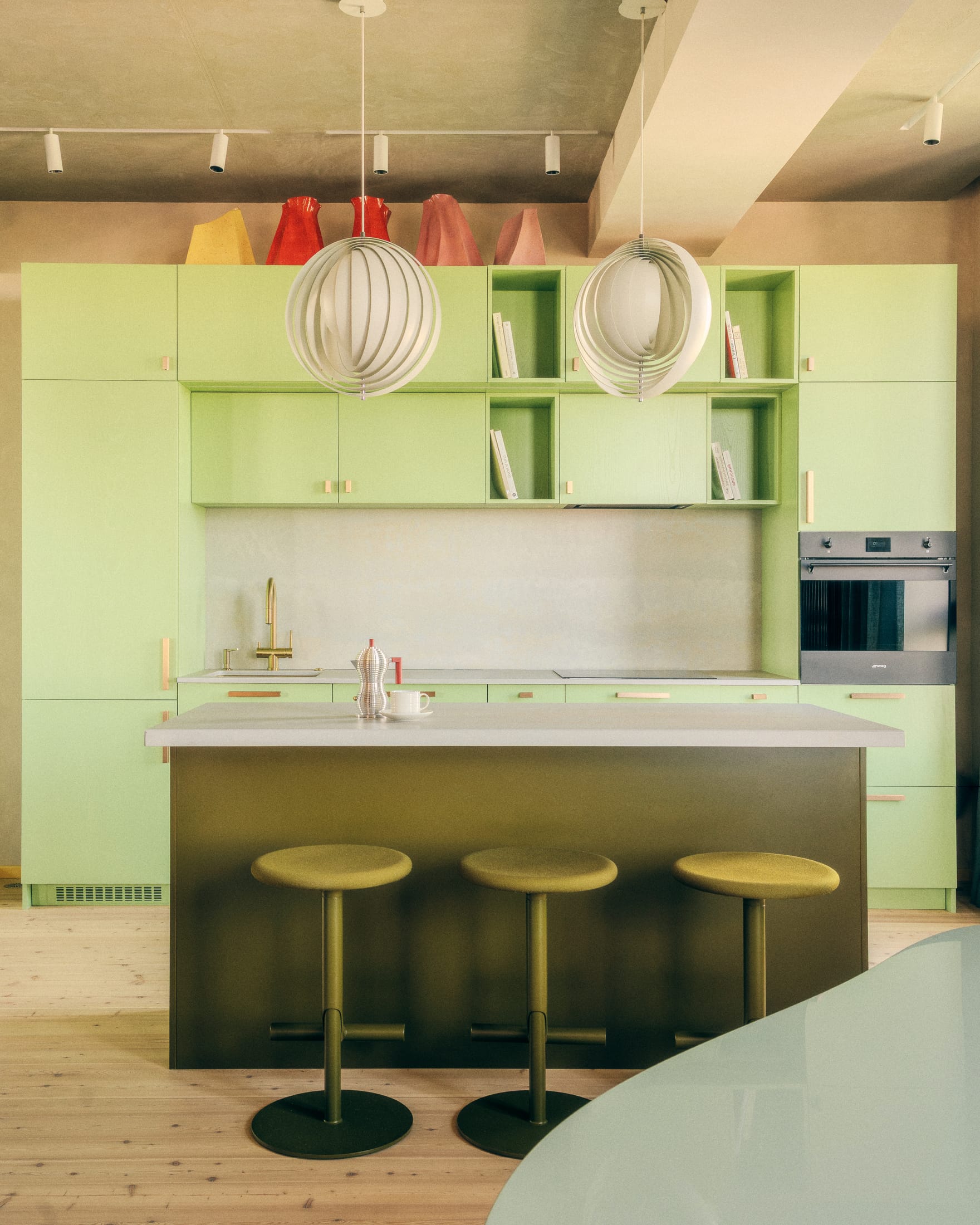 A modern kitchen with mint green cabinetry, a central island with olive green stools, and industrial pendant lighting.