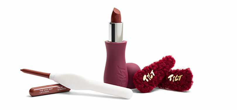 A red lipstick, matching liner, and fuzzy hair clips displayed on a clean white background.