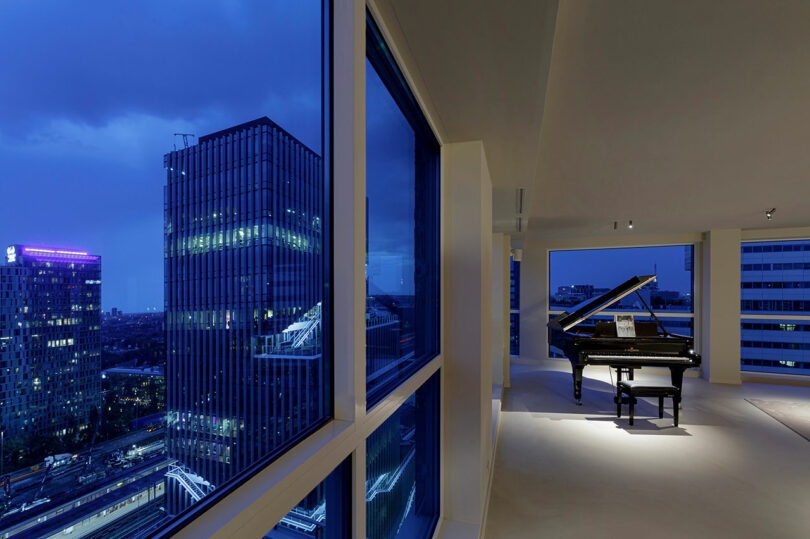 Modern minimalist living room with a grand piano and expansive floor-to-ceiling windows overlooking a night cityscape.