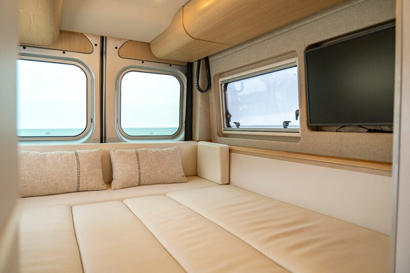 A cozy bedroom setup inside the camper van with a beige bed, wall-mounted TV, and a view of the ocean through the rear windows.