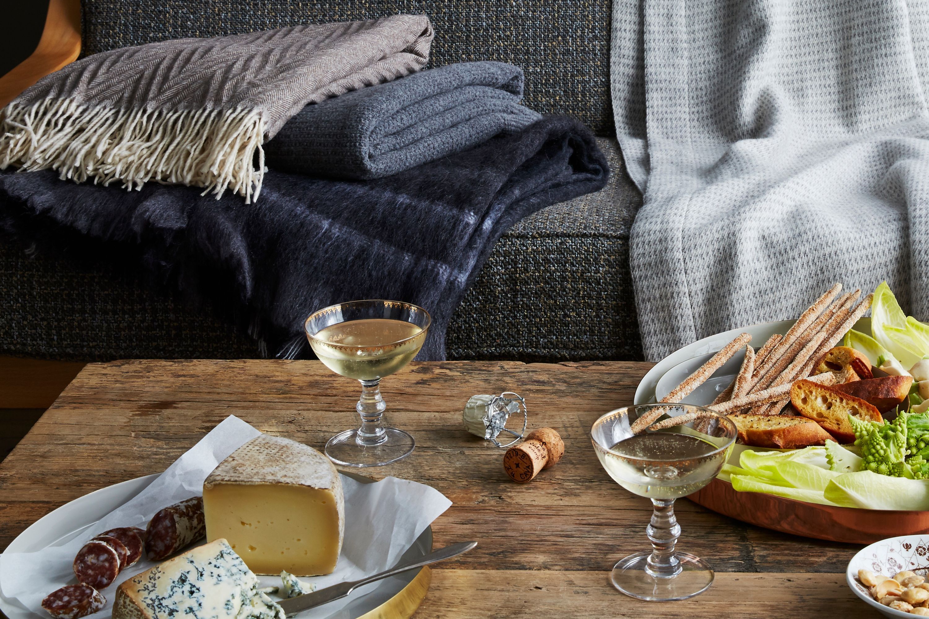 An inviting Scandinavian-style living room setup with a wine and cheese board, wooden furniture, and layered blankets.