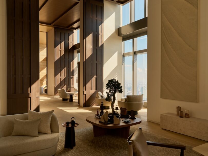 A vast modern living room with high ceilings, beige furniture, and sunlight streaming through large windows.