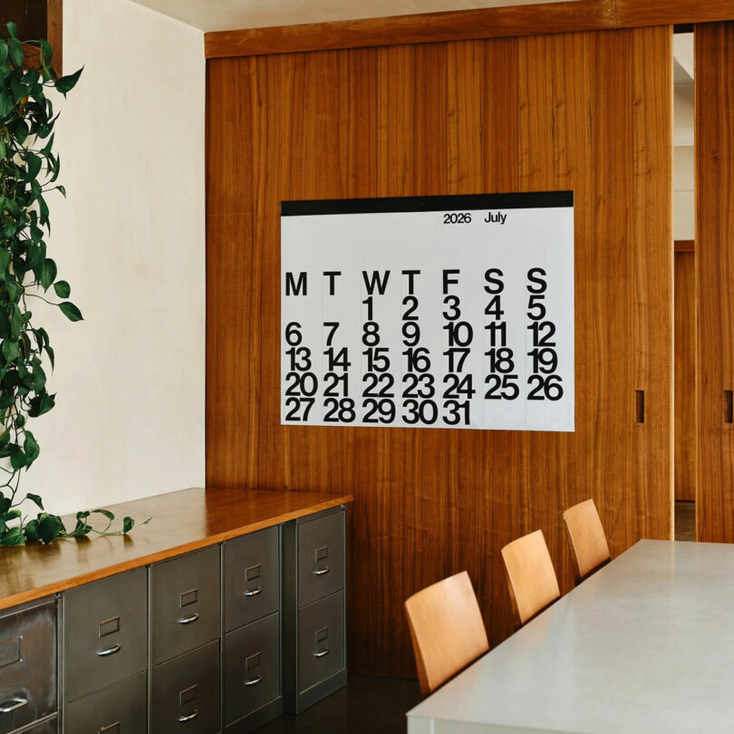 A large-format wall calendar displaying July in a modern office with metal cabinets and wooden furniture.
