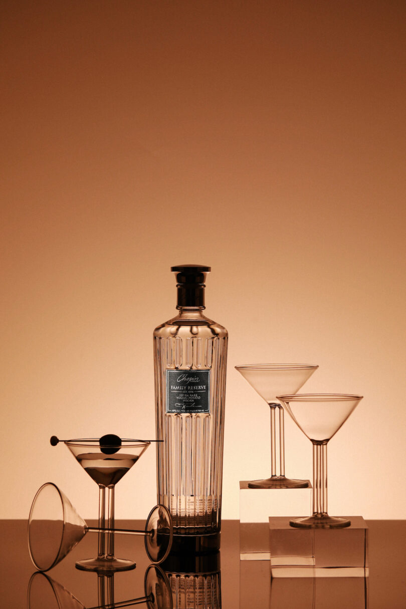 A clear glass bottle of Chopin vodka next to a filled martini glass with an olive against a warm orange gradient background.