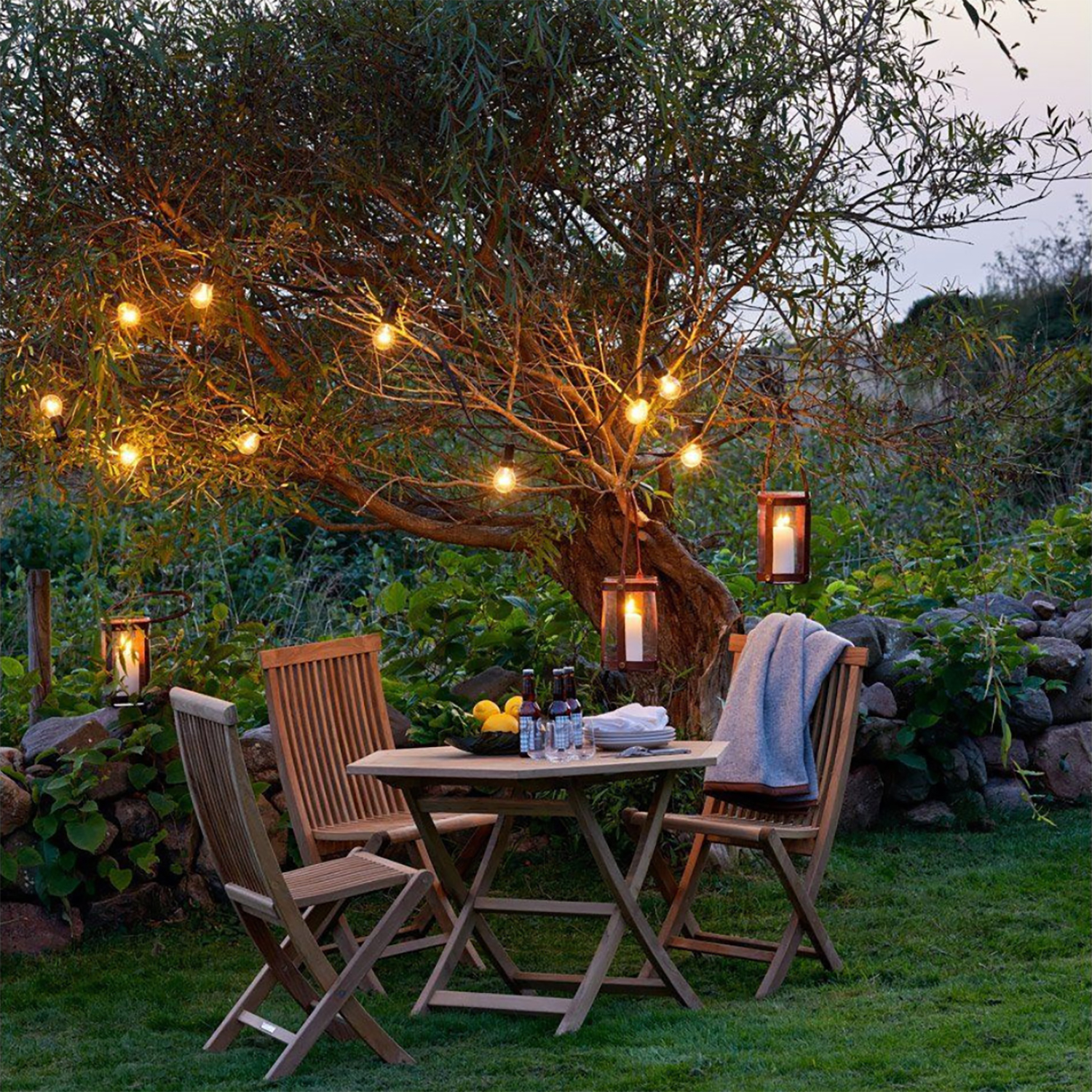 Cozy outdoor seating area under a tree adorned with warm string lights and candle lanterns surrounded by greenery.