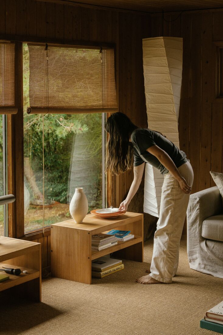 A woman sanding and moving pine tabletop elements in a sunlit room with pine walls.