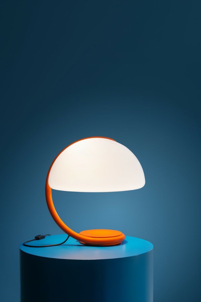A Serpente table lamp with an orange base and white shade set against a deep blue background.