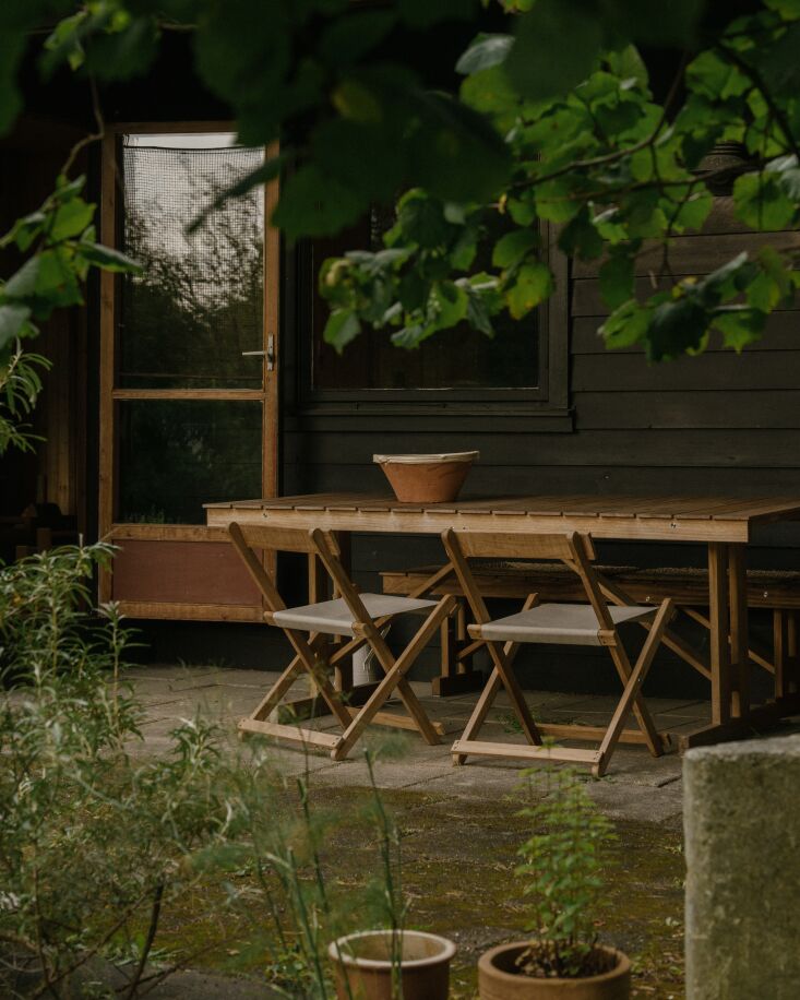 Outdoor dining set on a wooden terrace surrounded by flowers and trees with no visible fences.