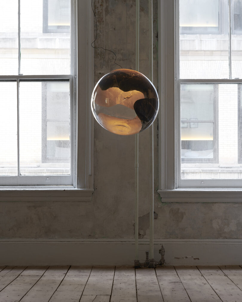 A single reflective pendant light suspended in a room with textured, peeling walls and large windows.