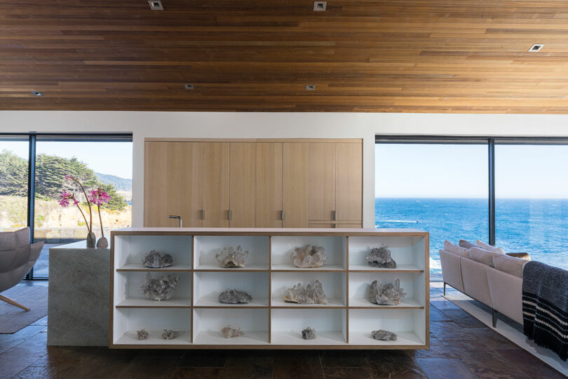 A living area with open shelving displaying organic shapes and sea-inspired decor against a coastal backdrop.