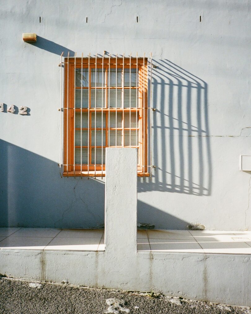A barred window casting a sharp linear shadow on a pale blue wall with an orange frame.