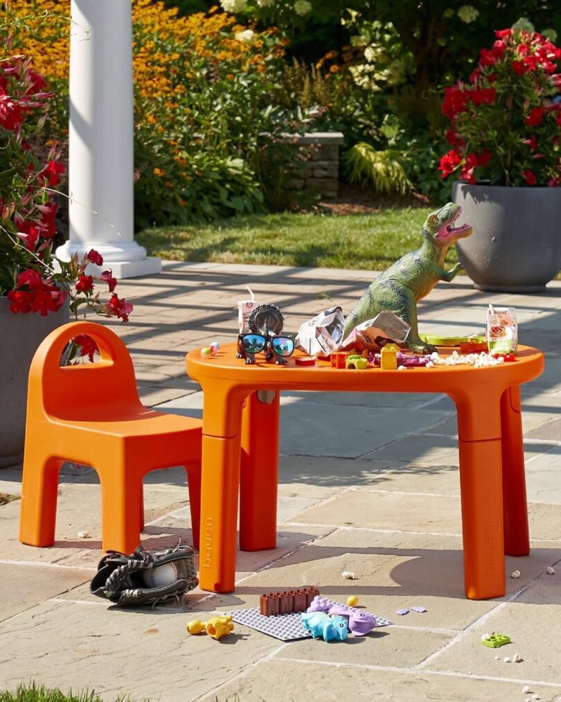 A modern orange children's table and chair set placed outdoors among red flowers and toys.