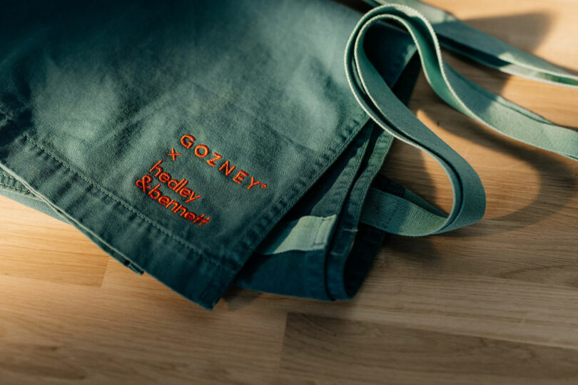A green Gozney x Hedley & Bennett branded tote bag on a wooden surface.