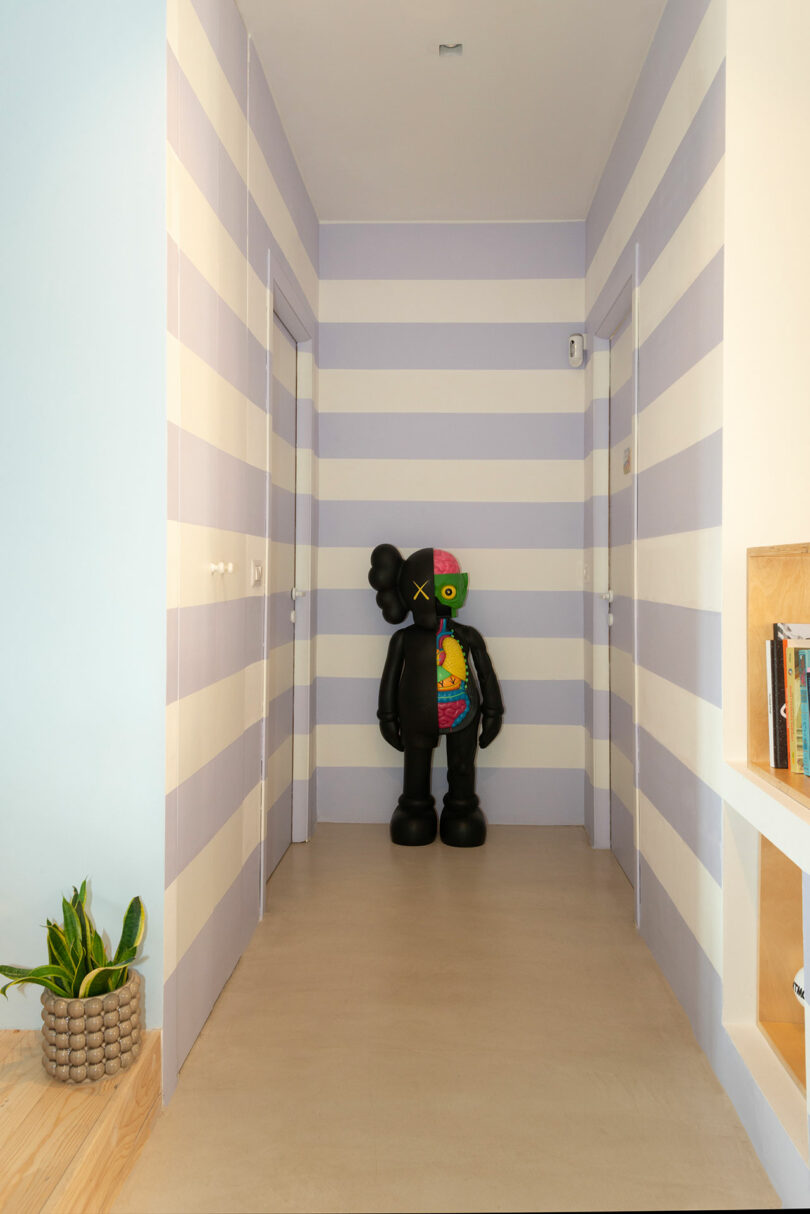 A modern hallway with horizontal lilac and white stripes featuring a black KAWS sculpture.