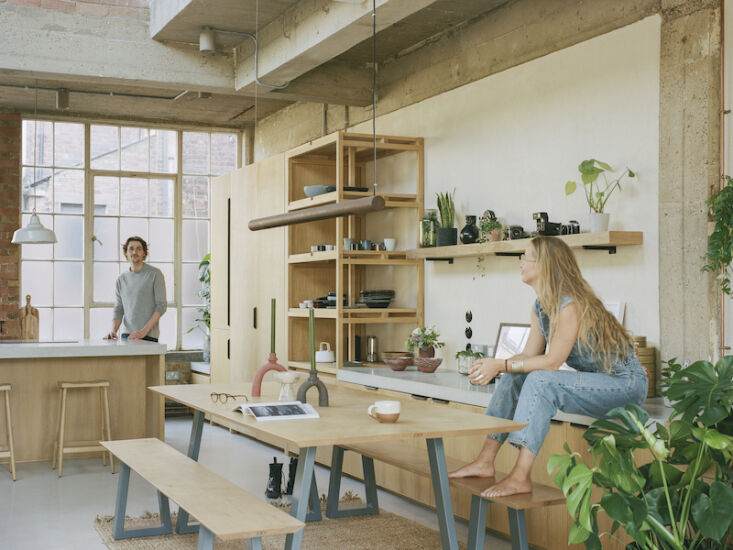 Two people working and interacting in a modern industrial kitchen and living area with minimalist decor.