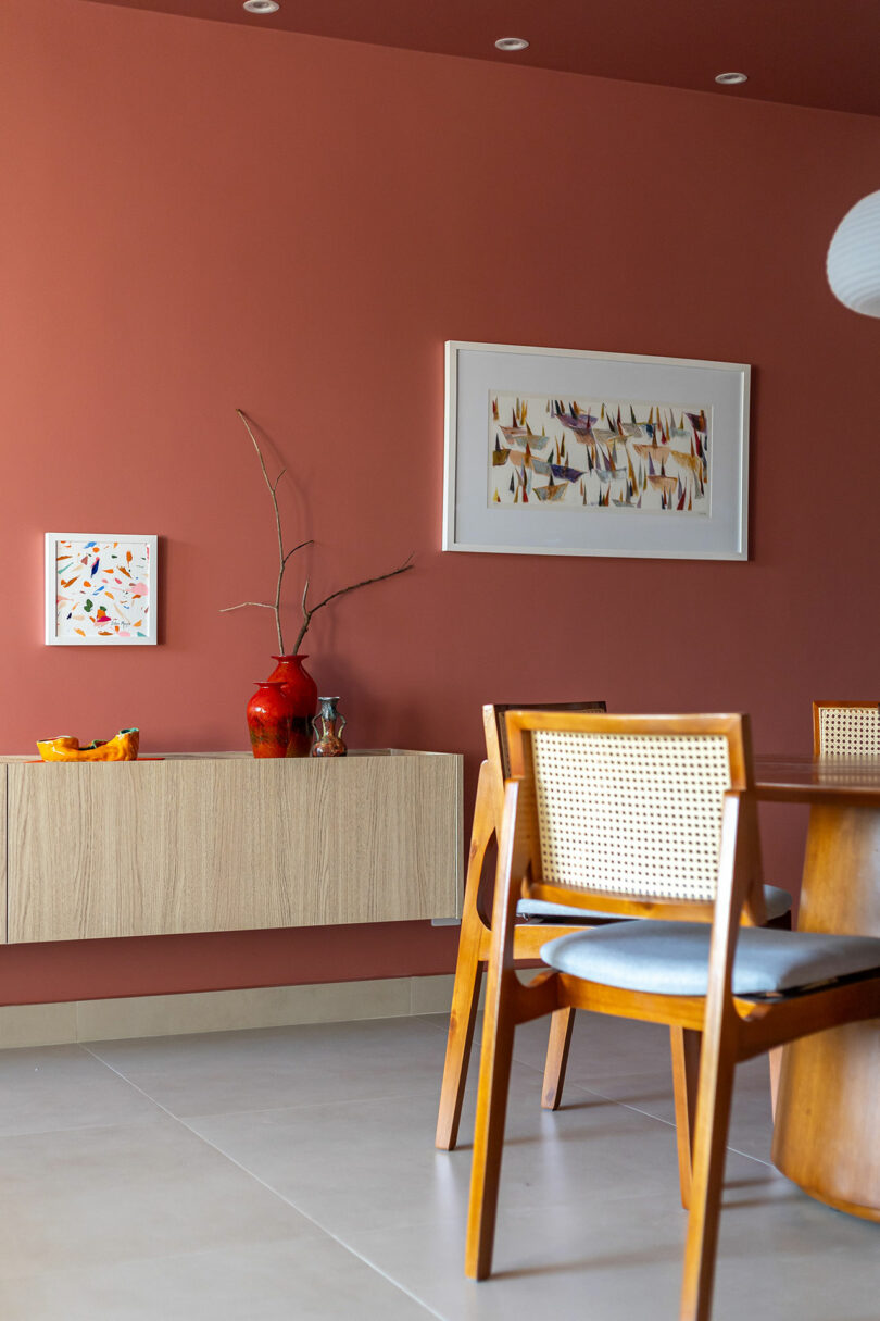 Modern dining area with a floating wooden sideboard, decorative vases, and framed art against a terracotta wall.