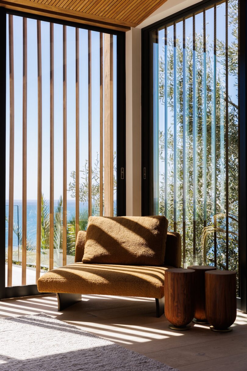 A brown armchair by floor-to-ceiling windows with vertical wooden slats casting shadows.