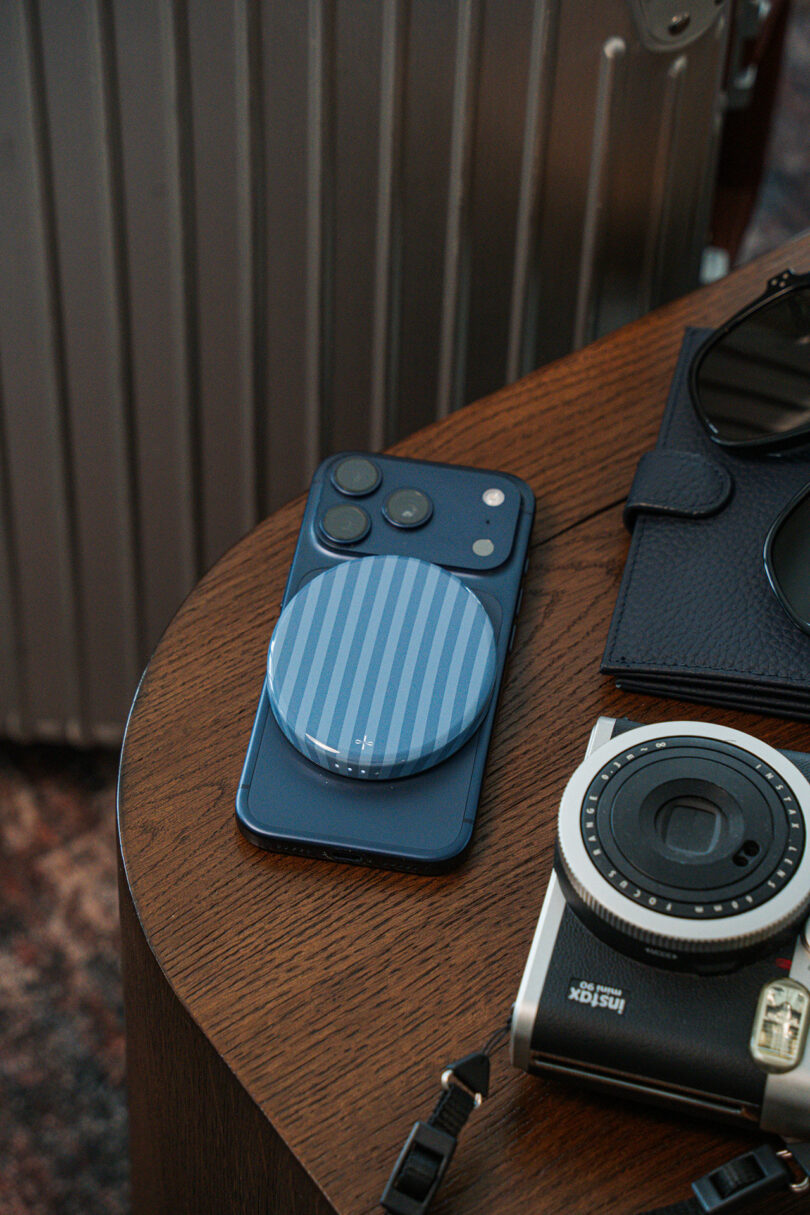 A blue smartphone with a striped charger on a wooden table alongside a camera and sunglasses.