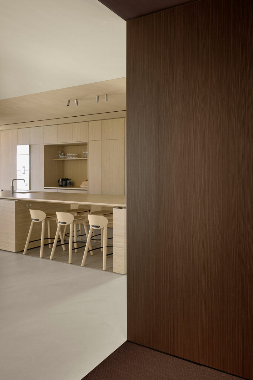 Modern minimalist kitchen with light wood cabinetry, a long island with stools, and dark wood panels.