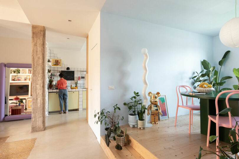 An open-plan living and dining area with light blue walls, a green dining table, and a person standing in the background kitchen.