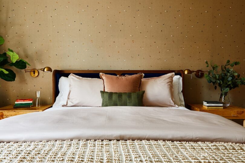 Close-up of a bed with neutral linens against a backdrop of dotted beige Dedar wallpaper.
