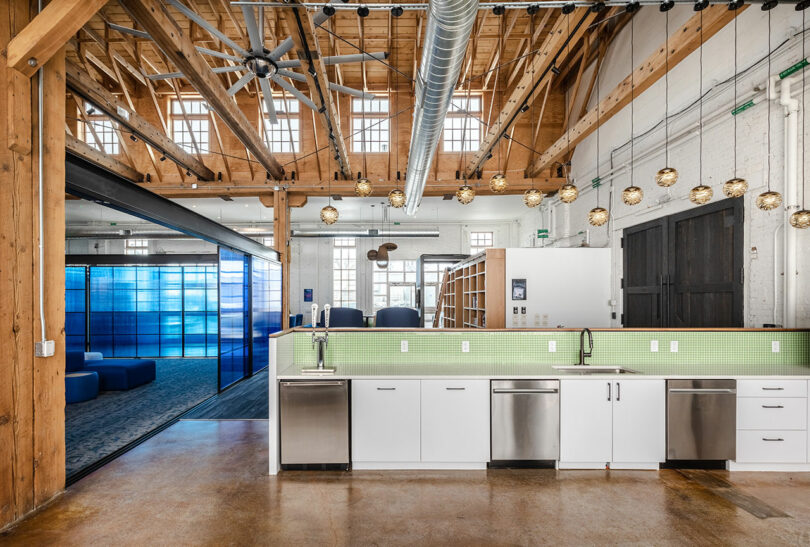 A modern office kitchen with green tile backsplash and white cabinets adjacent to an open workspace.