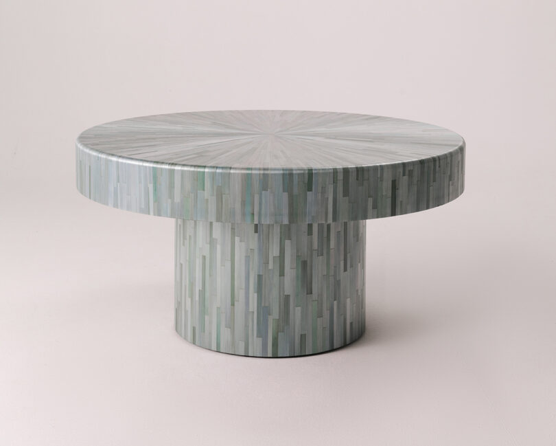 A round coffee table featuring light green and gray straw marquetry with a thick cylindrical base.