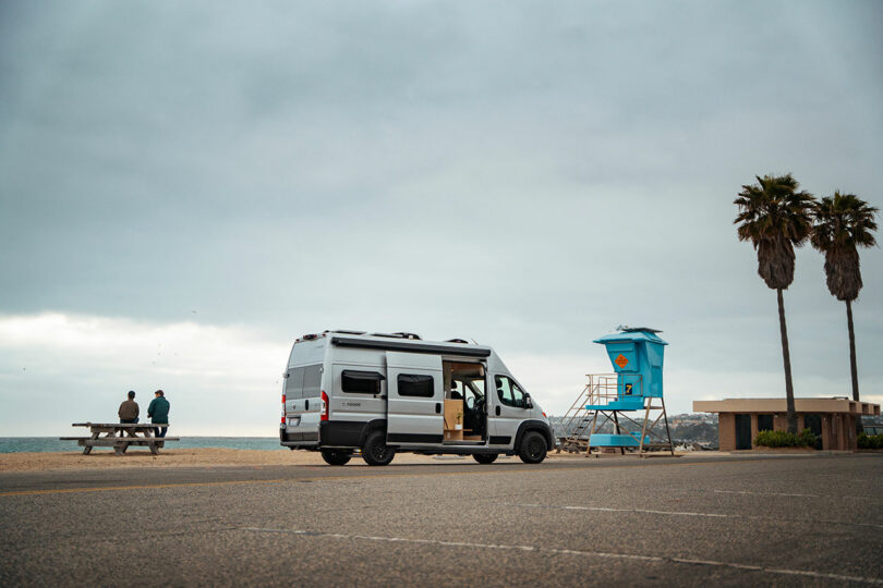 Silver Noovo camper van parked on a coastal road near palm trees and a lifeguard tower.