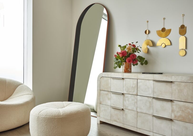 A serene bedroom corner with an arched mirror, cream-colored dresser, and a curved velvet chair, creating a peaceful retreat space.