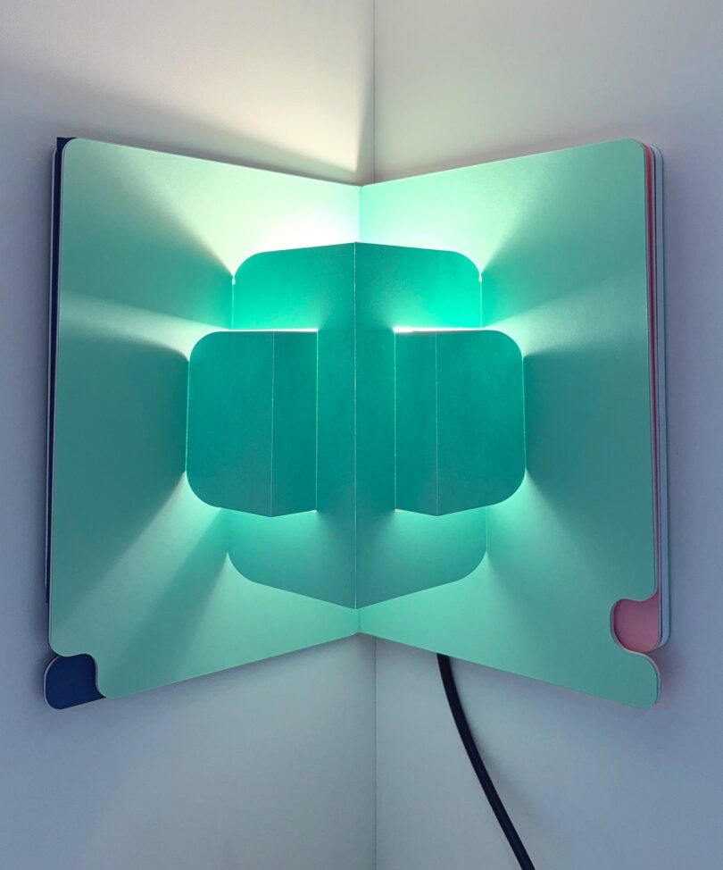 An open pop-up book lamp emitting a blue-green glow with geometric shadows in a room corner.