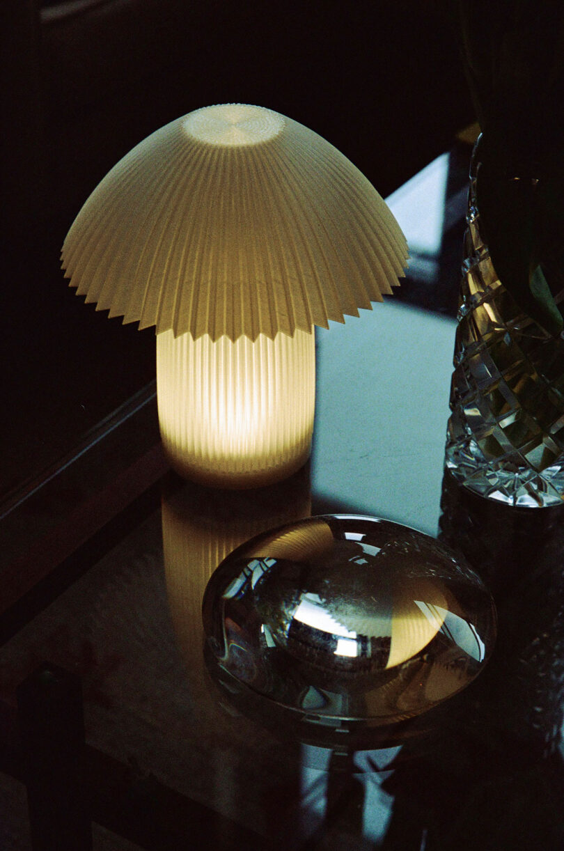 A pleated, mushroom-shaped warm glowing lamp on a glass table next to decorative glass objects.