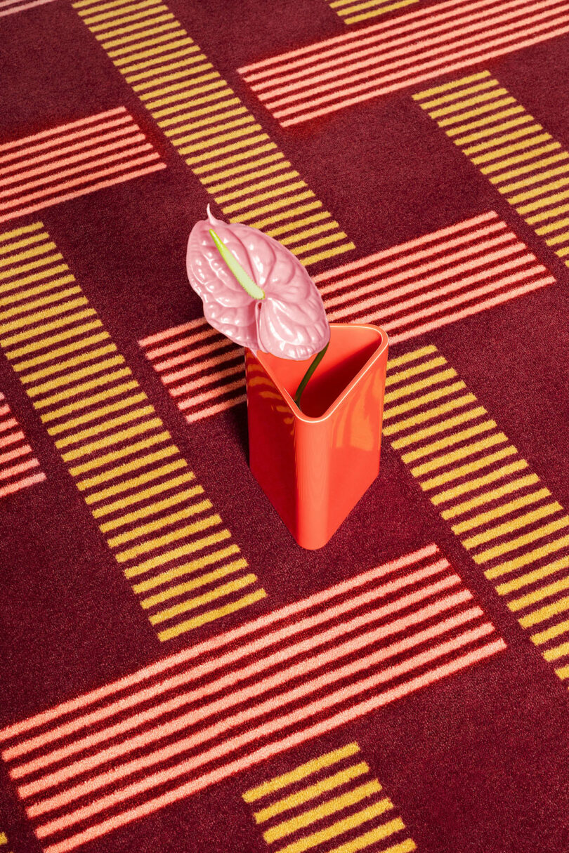 A red anthurium flower in an orange vase on a red and yellow striped geometric carpet.