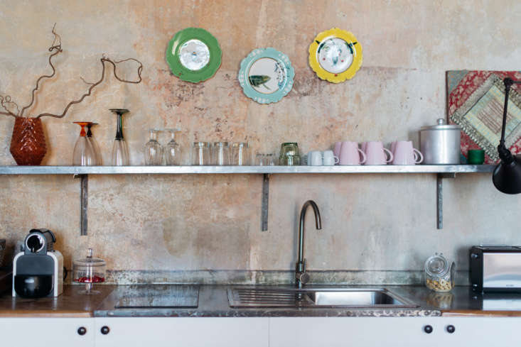 Rustic kitchen featuring textured plaster walls and industrial open shelving with colorful mugs.