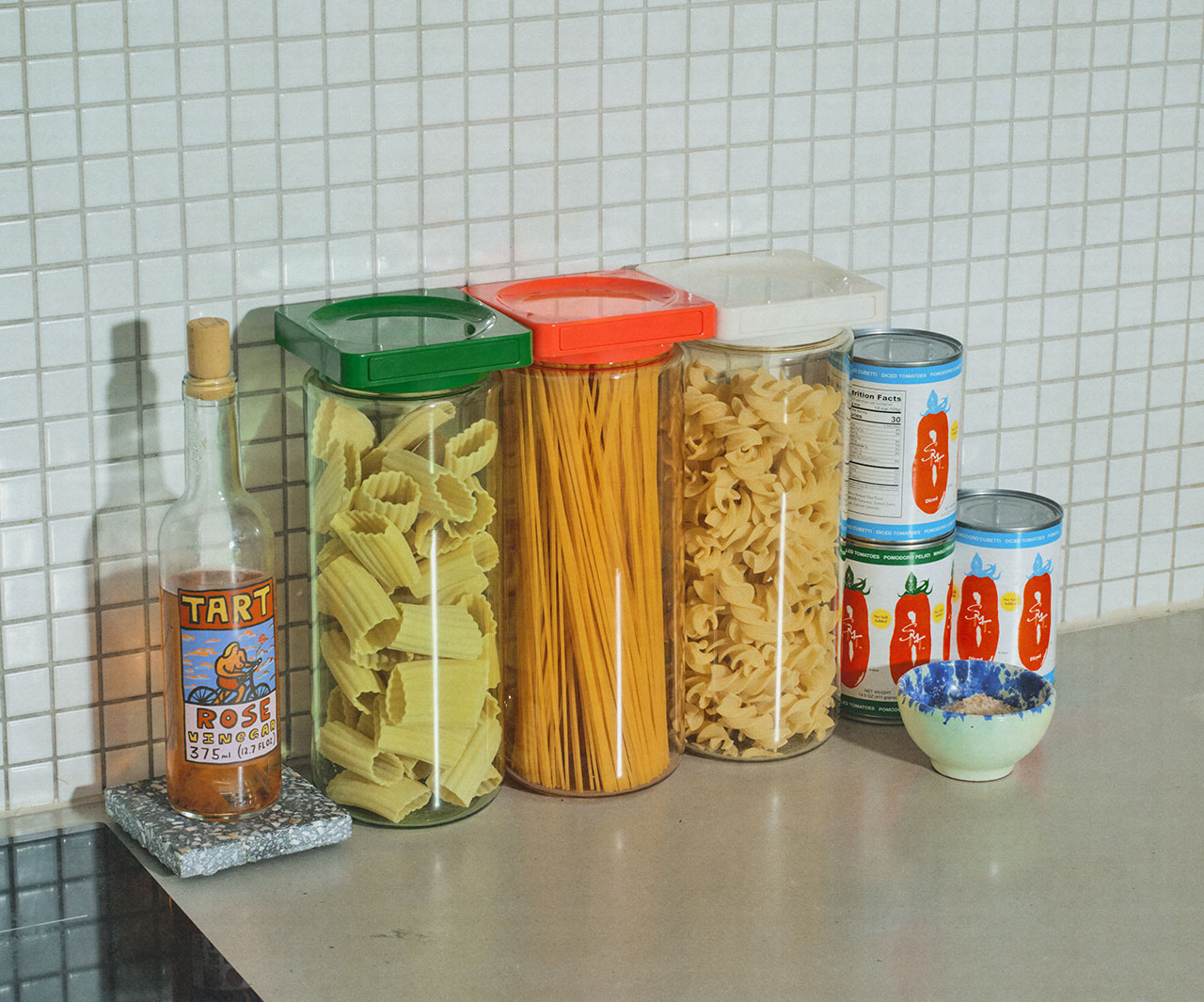 Kitchen countertop with colorful pasta containers, a bottle of rose vinegar, and canned tomatoes against a white tiled backsplash.