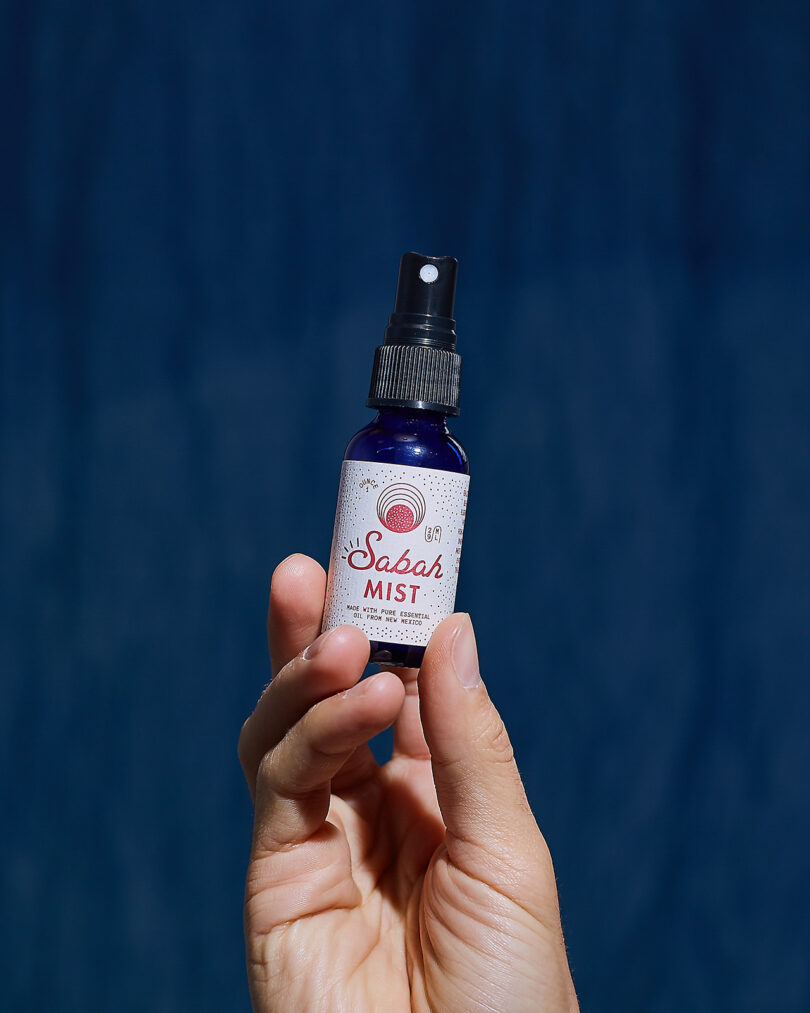 Hand holding a blue Sabah Mist spray bottle against a moody blue background.