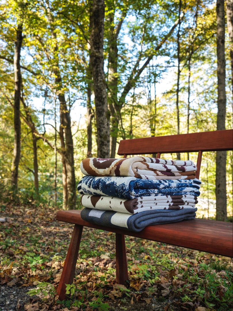 A stack of folded blankets on a wooden bench in a sun-dappled forest setting.