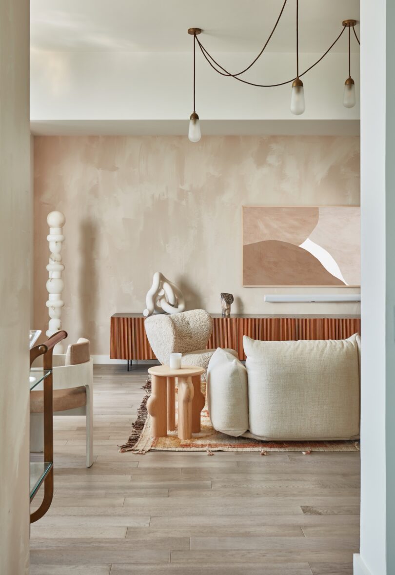 A modern living room with neutral-toned textured furniture, sculptural decor, and light wood flooring under soft pendant lighting.