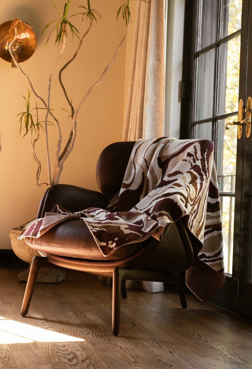 A patterned blanket draped over a brown armchair in a bright, sun-filled room with a large window.