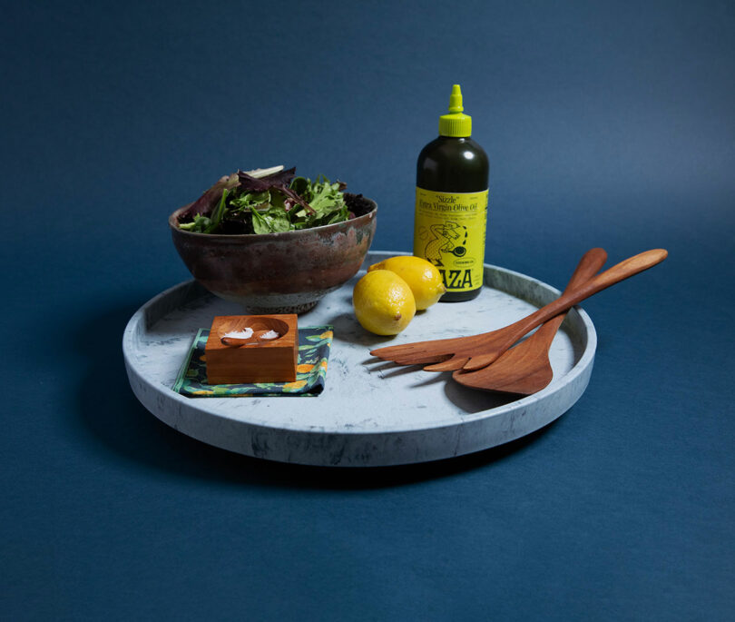 A marble and wood turntable (Lazy Susan) styled with lemons, greens, and kitchen accessories against a blue background.